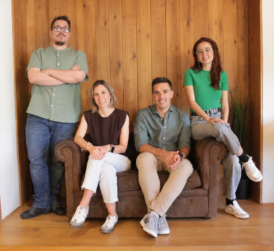 El equipo de Normatia en la oficina: Alfonso de pie a la izquierda, Verónica y Ricardo sentados en un sofá Chester y María sentada en el brazo del sofá, ante una pared de madera.